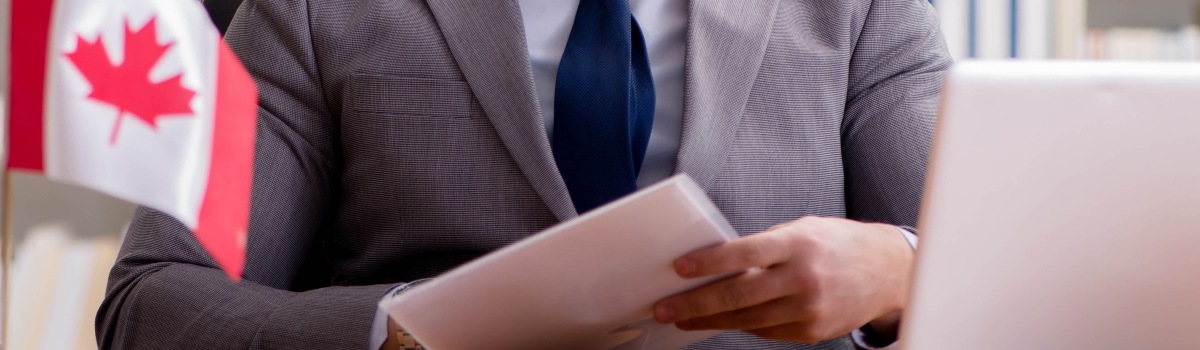 A businessman sits at a desk and reviews paperwork. A Canadian flag, computer, and notebook are on the desk. 