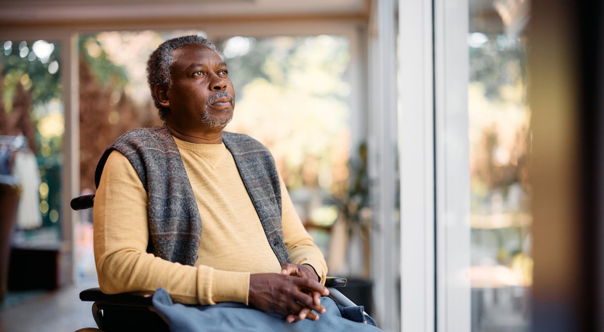 An aged man in a wheelchair sits outside and gazes despondently upwards.