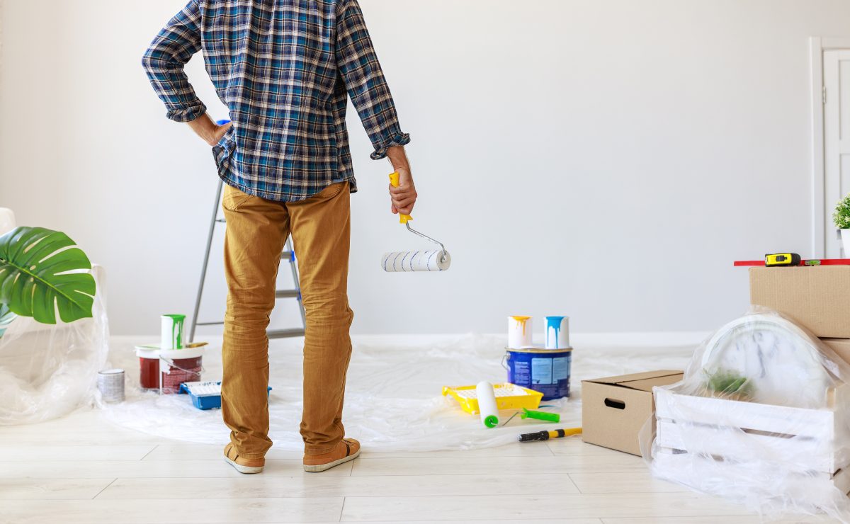 A man stands in a mostly empty room holding a paint roller. He is surrounded by cans of paint, tarps, moving boxes, and a ladder.