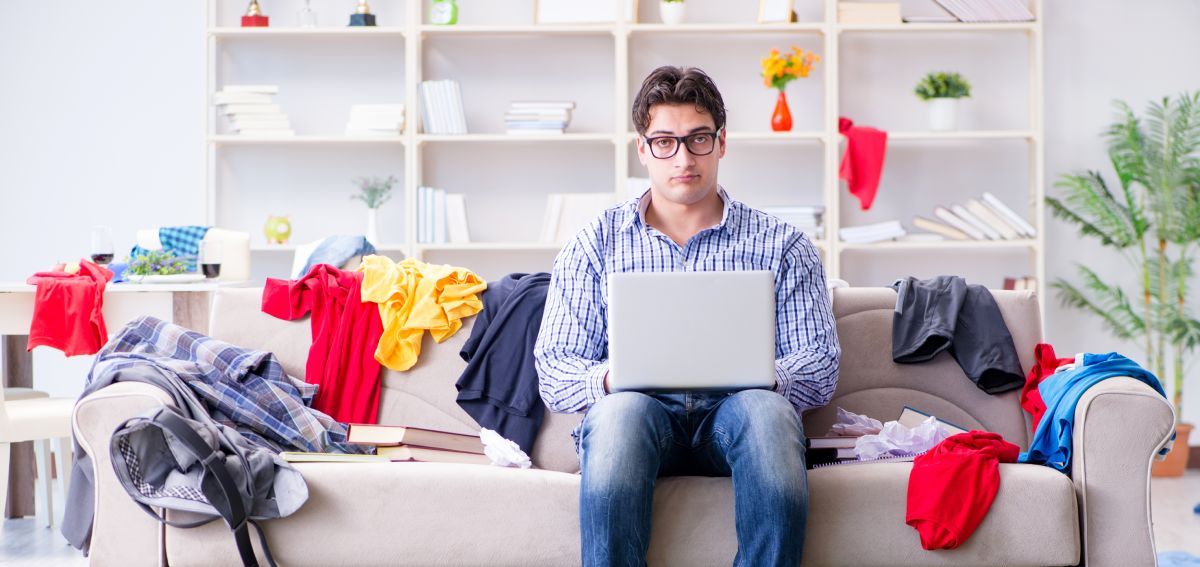 A man sits on a couch with a laptop. He has an unsatisfactory look on his face and is surrounded by heaps of clothes, books, and trash.