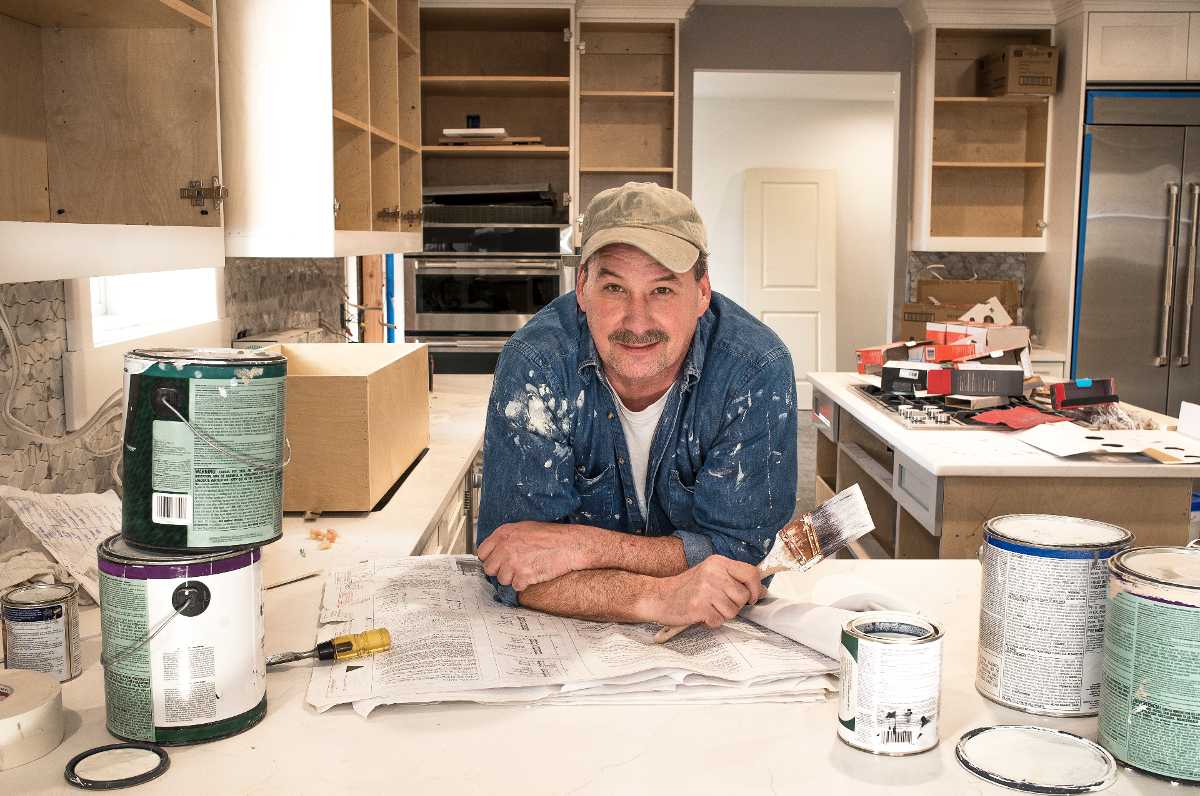 A man holds a paintbrush and leans over a counter in a kitchen that's in the middle of extensive renovations.