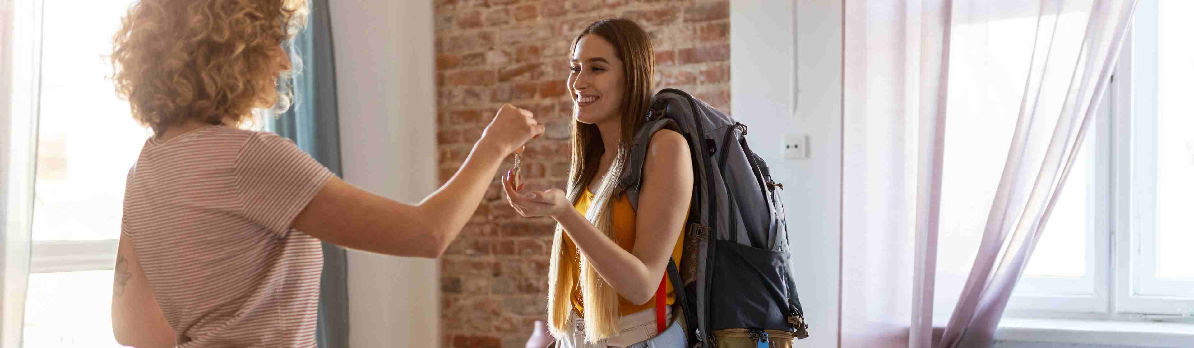 A young female backpacker renting an apartment. She is receiving a set of keys from another young woman. They are both smiling.