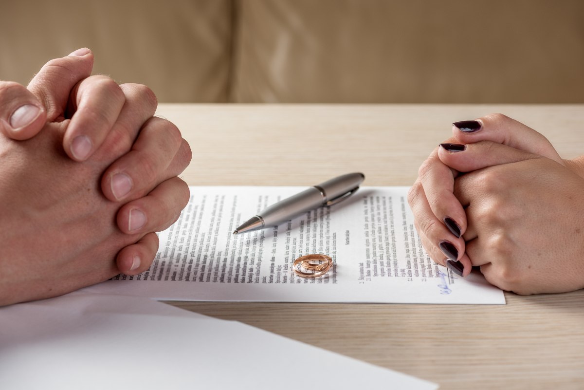 Two people place their wedding bands on a stack of divorce papers on a table between them. 