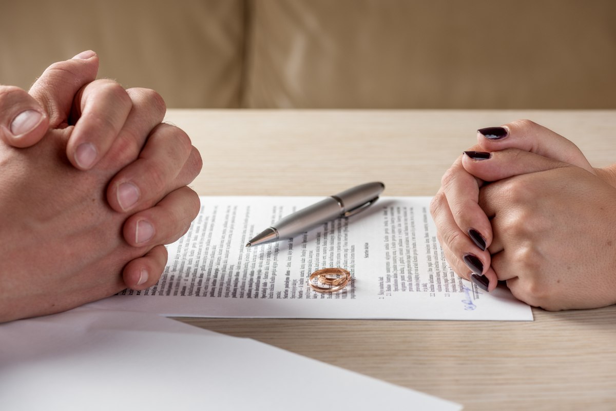 Two people place their wedding bands on a stack of divorce papers on a table between them.
