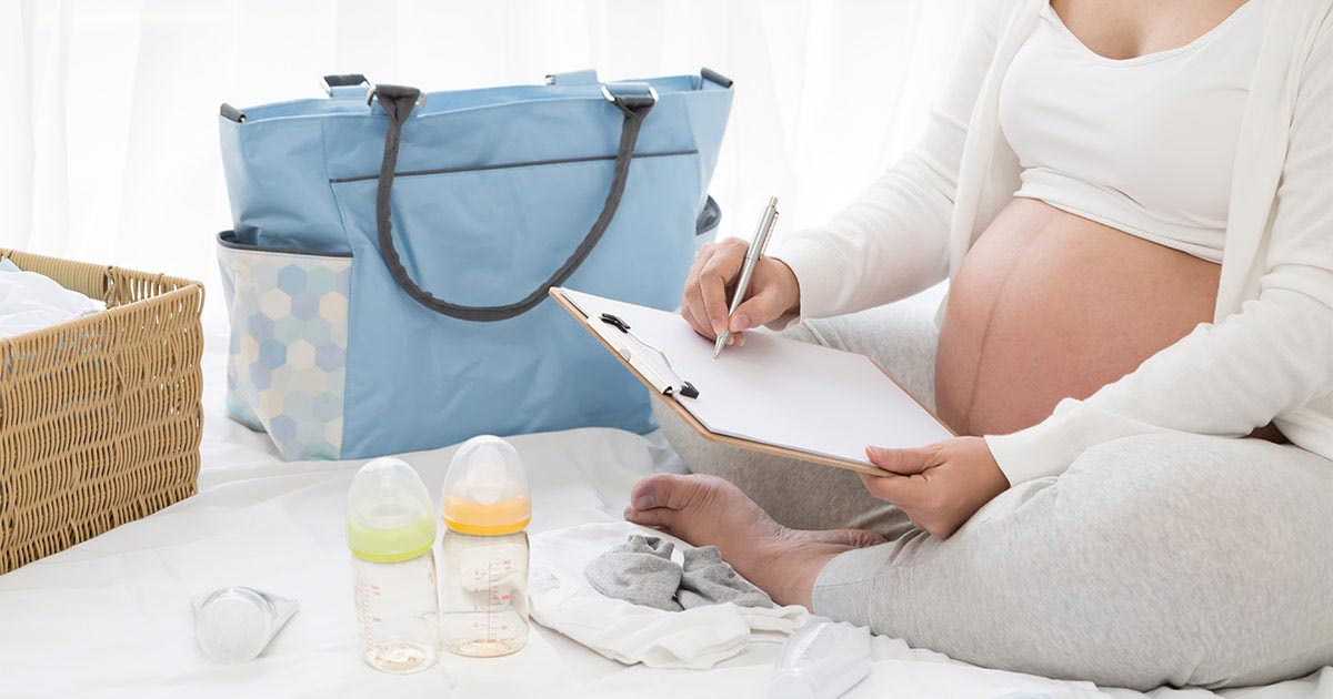 A woman in her third trimester of pregnancy sits cross-legged on a bed, holding a clipboard and pen. She has baby bottles, clothes, and a diaper bag nearby.