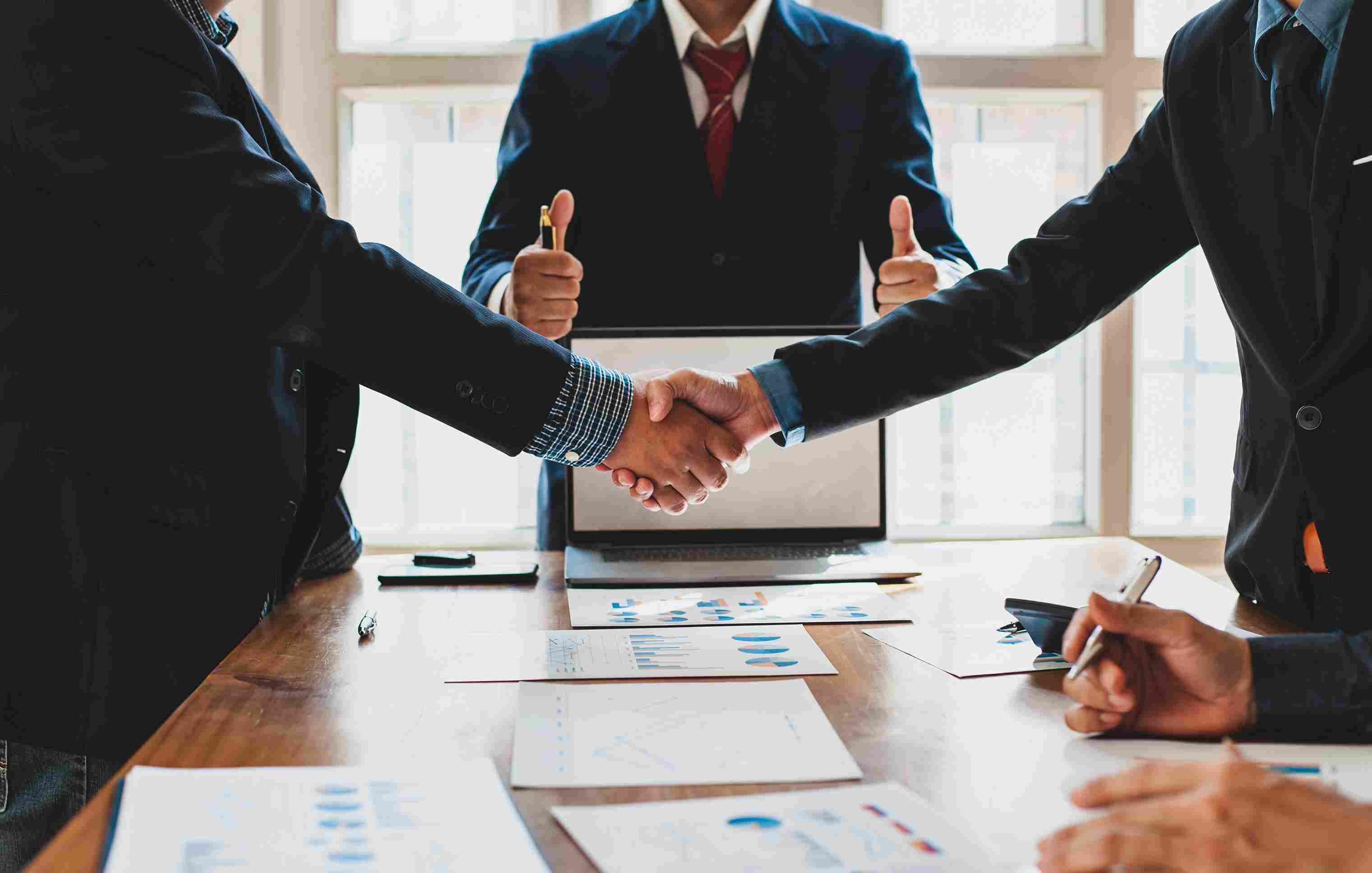 Two people in suits shaking hands over a boardroom table with another person holding two thumbs up behind them