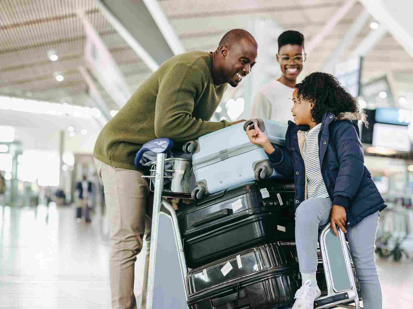 A father and his daughters hold hands walking through an airport. The father is wheeling a carry-on sized suitcase with his left hand and holding the older daughter's hand with his right. The older daughter is holding the youngest's hand.