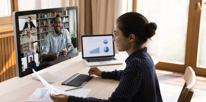 A woman joins a video conference call from her home office. She smiles back at the screen, which shows several of her co-workers smiling, talking, and taking notes.