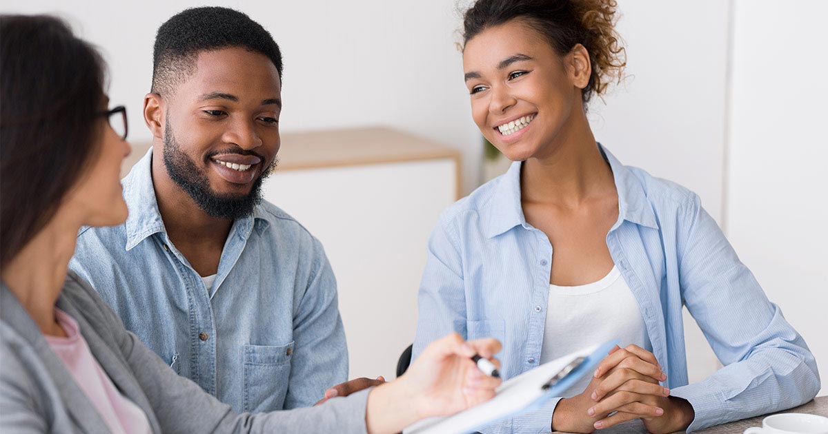 A young, heterosexual couple smiles as they sit and talk with a professional person at a table.