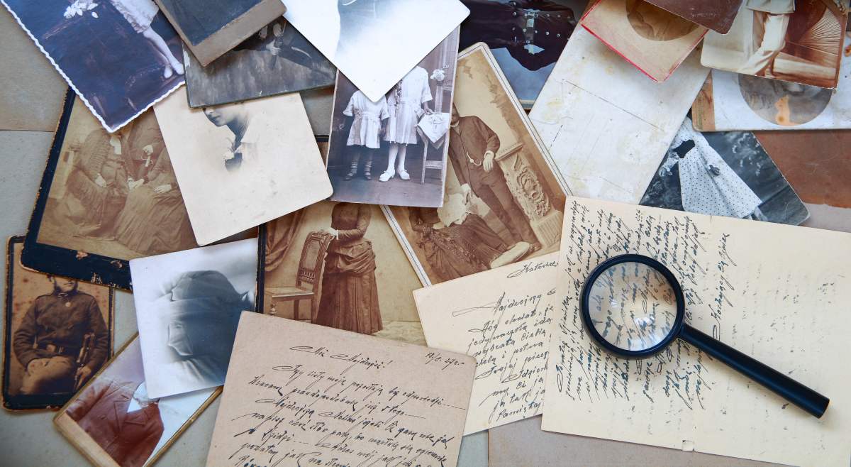 A magnifying glass rests on top of a pile of vintage photographs and handwritten letters that are scattered across a table. 