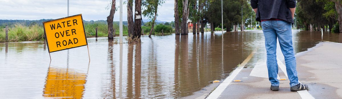 With his back to the camera, a man stands at the edge of a road that is impassible because it is flooded with water. There is a yellow square sign jutting out from the water that reads WATER OVER ROAD.