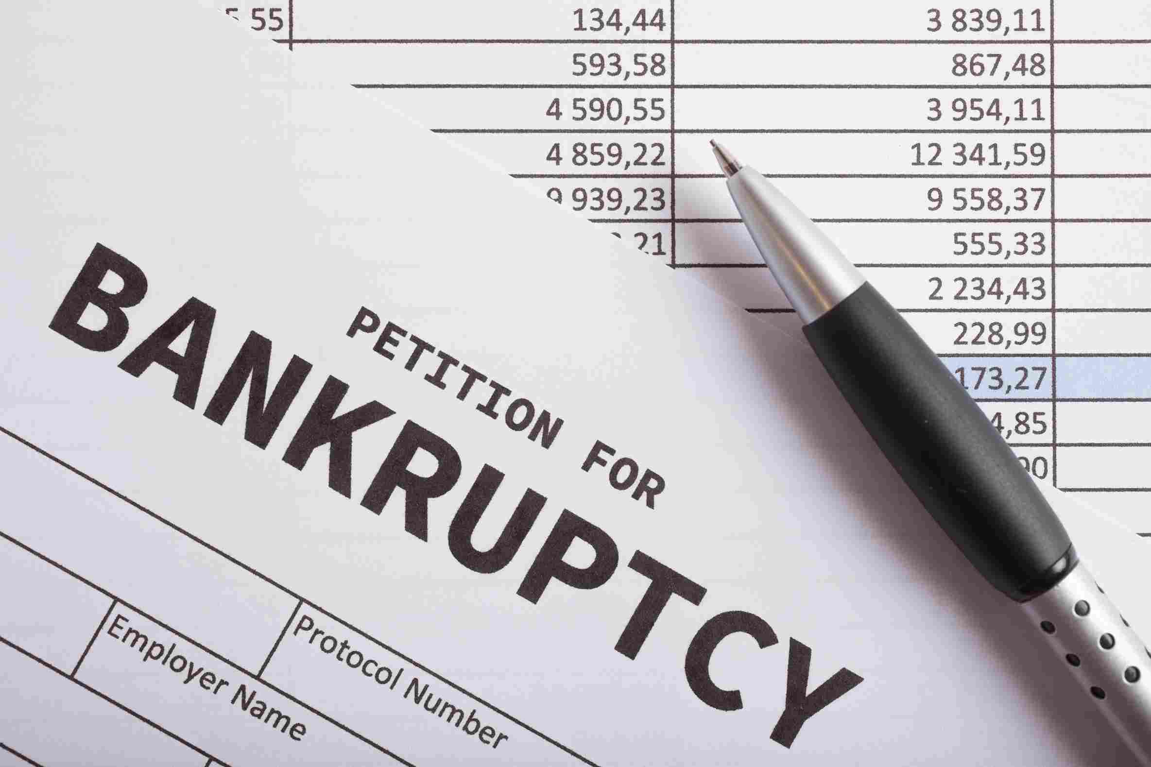 The words petition for bankruptcy are in bold black print on a stack of papers and spreadsheets. A ballpoint pen rests on top.