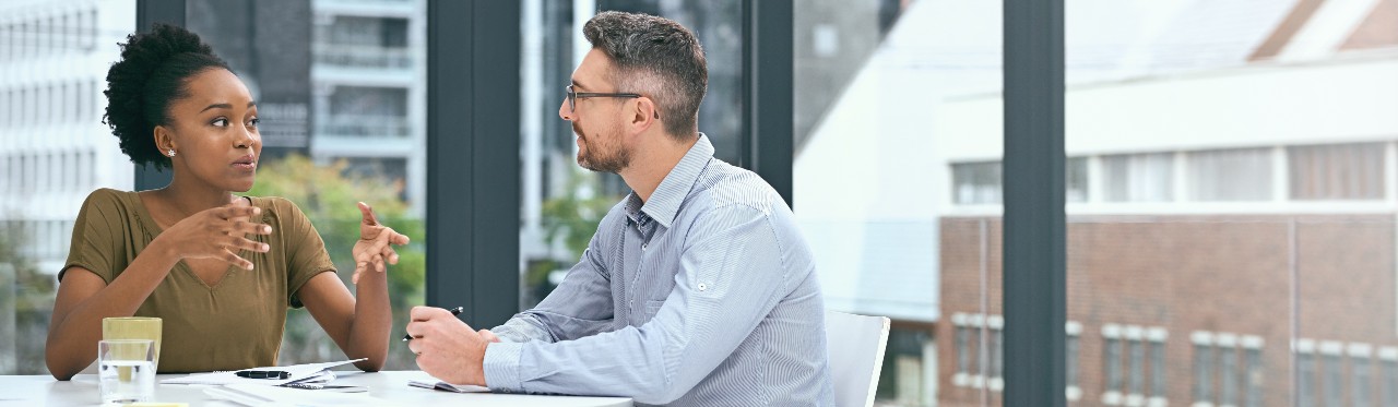 A man and a woman sit at a table in a room with many windows. The woman is speaking and the man is listening. 