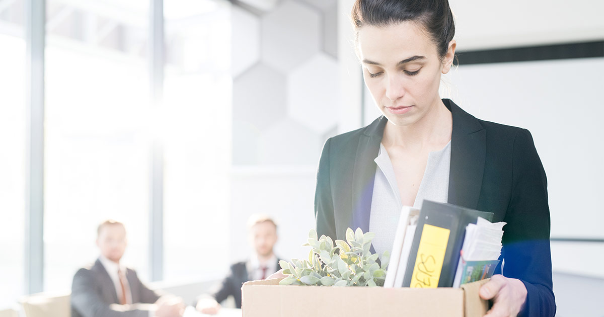 A woman walks out of an office carrying a box of office supplies and plants. Her eyes are downcast, and she has a solemn expression.