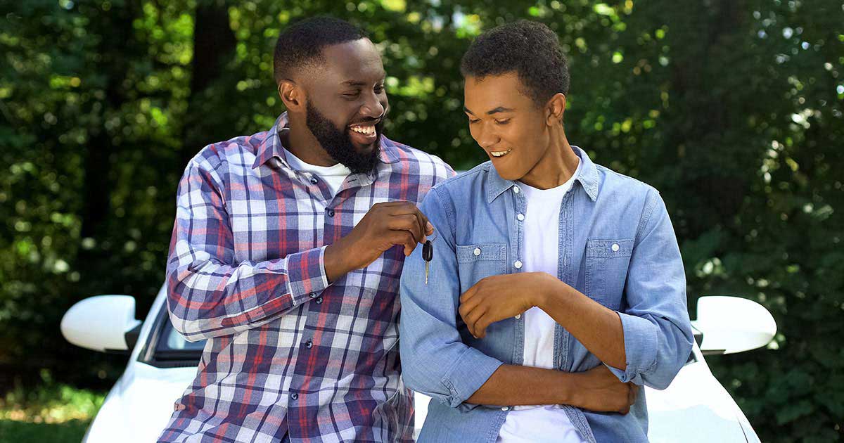 A man smiles and hands a key to an excited teenager as they sit on the hood of a white car.