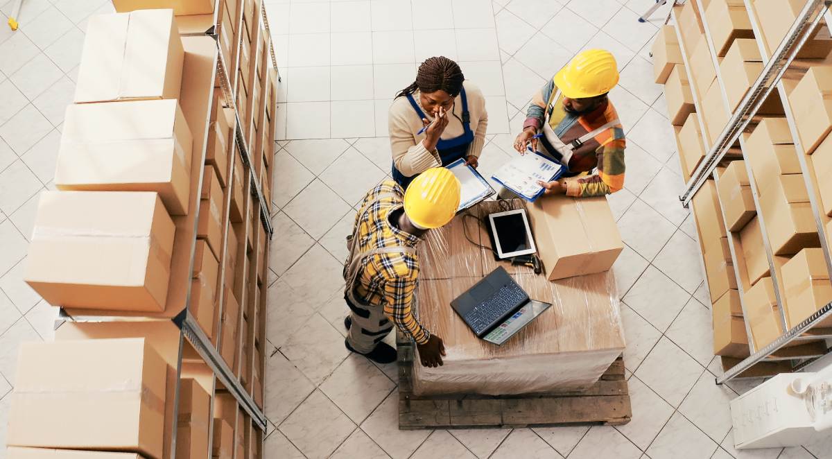 Three workers wearing coveralls and hard hats look at paperwork while standing in the middle of a warehouse filled with neatly stacked cardboard boxes.