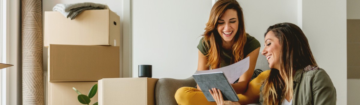 two women smiling while looking at a tablet and paperwork with unpacked moving boxes behind them