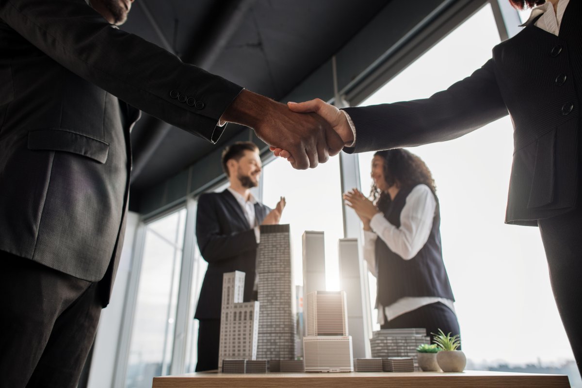 A group of business people stand around a diorama of a building complex; they shake hands and celebrate.