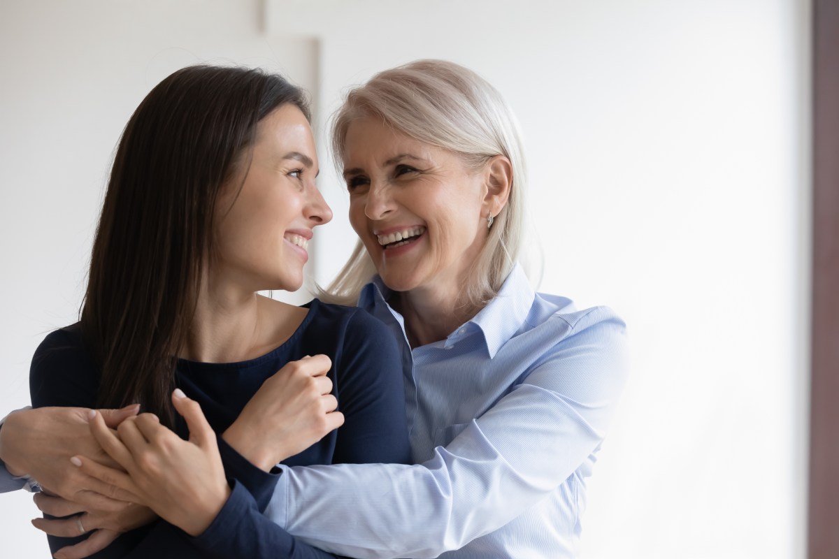 An elderly mother hugs her adult daughter from behind. They're both laughing.