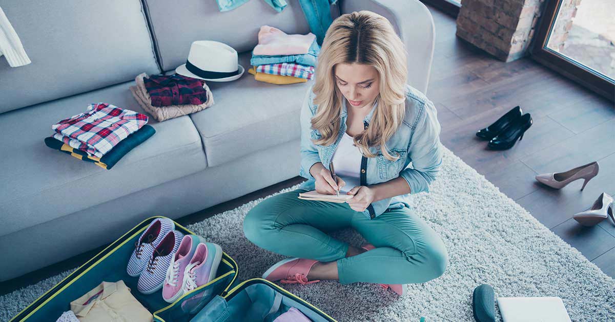 A woman sits cross-legged on her living room floor, looking down at the notebook and pen in her hands. She has an open suitcase in front of her, half packed with clothes and shoes. More clothing and shoes are spread out on the couch and floor around her.