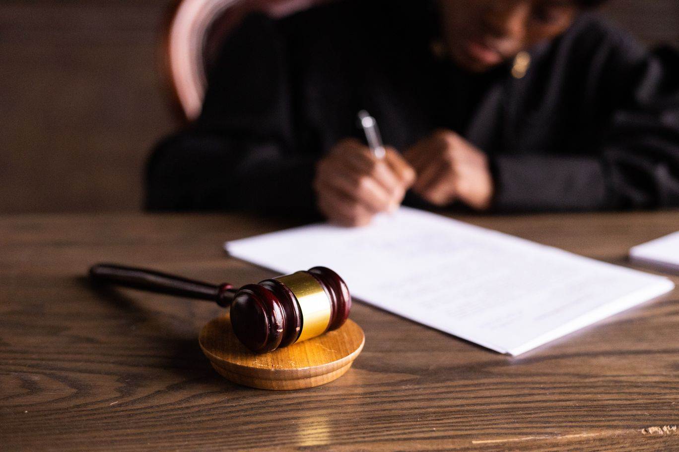 A person in black judge's robes sits at a desk and signs papers. The focus is on a gavel that sits on the desk. 