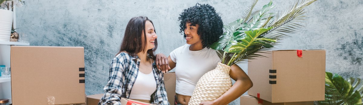 Two happy, smiling women stand close to each other. They are surrounded by cardboard boxes.