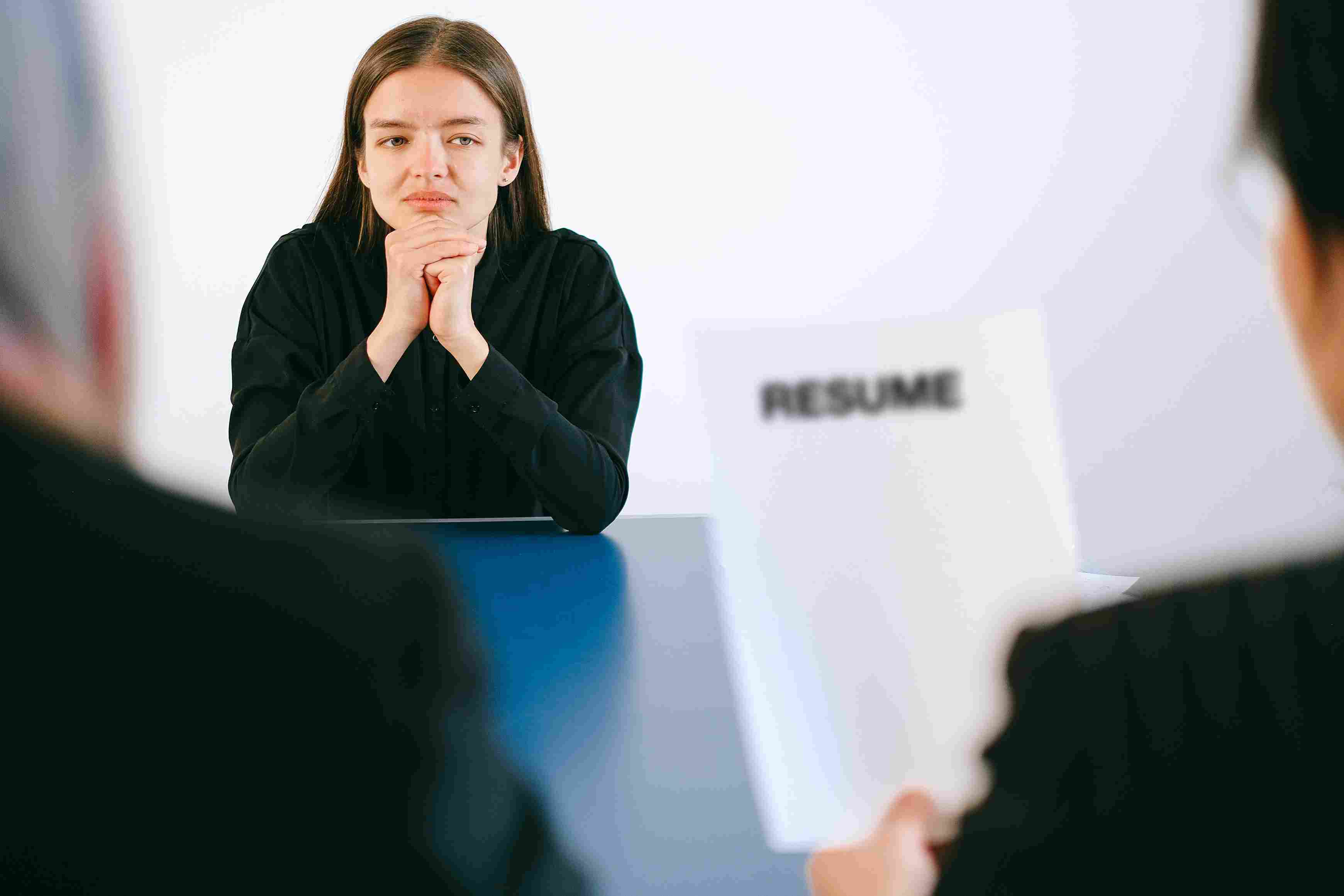 A pair of hands are folded. The person's face is out-of-frame. Another out-of-frame person holds a paper that says RESUME.