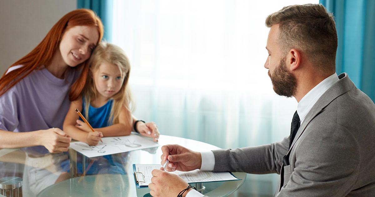 A mother sits at a table with her daughter in her lap. The child looks up from her drawing at a professional-looking man across from her.