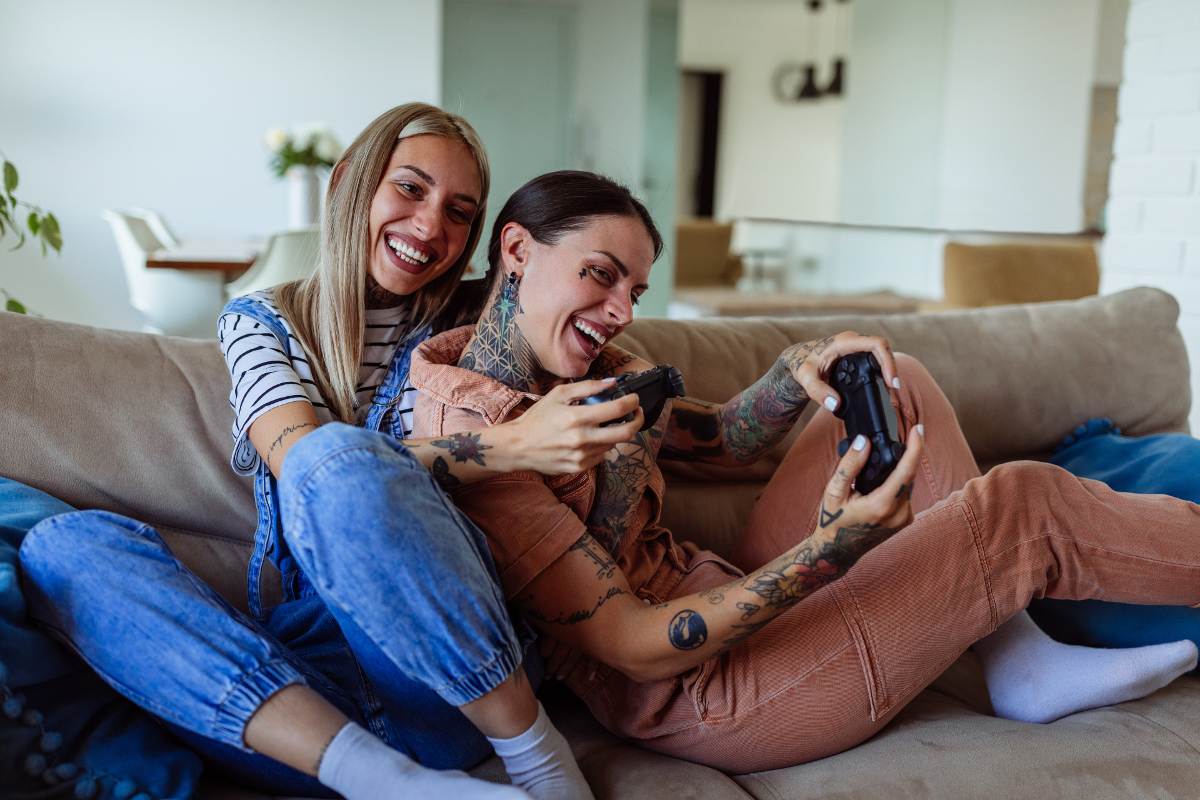 A lesbian couple cuddles on a couch while laughing and playing video games. 