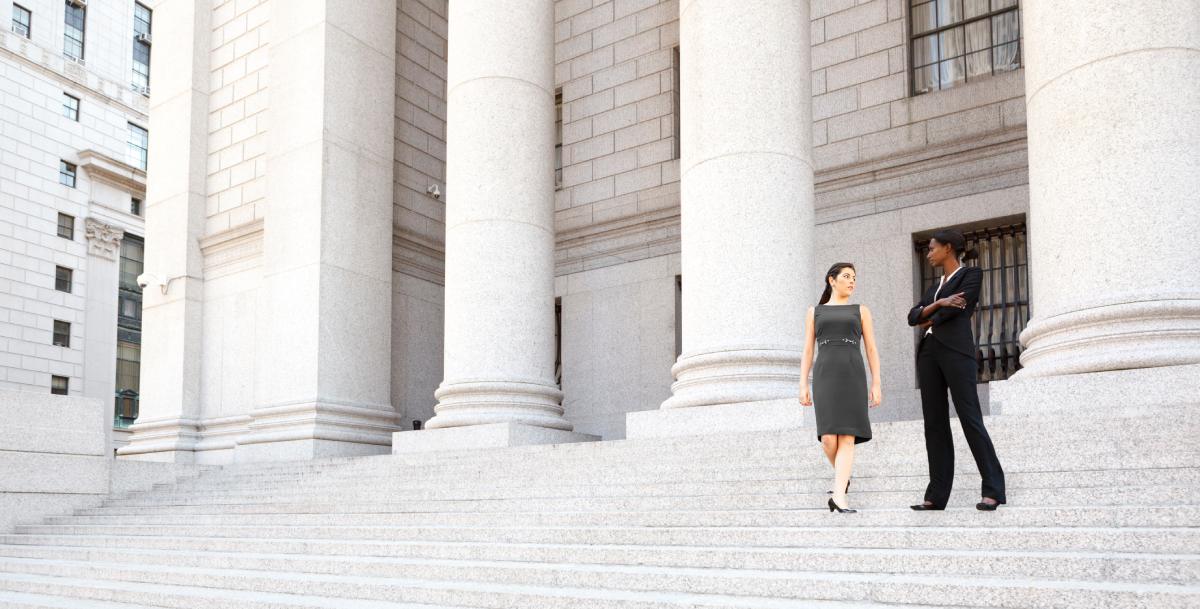 Two professionally dressed women stand on the steps outside of a courthouse.