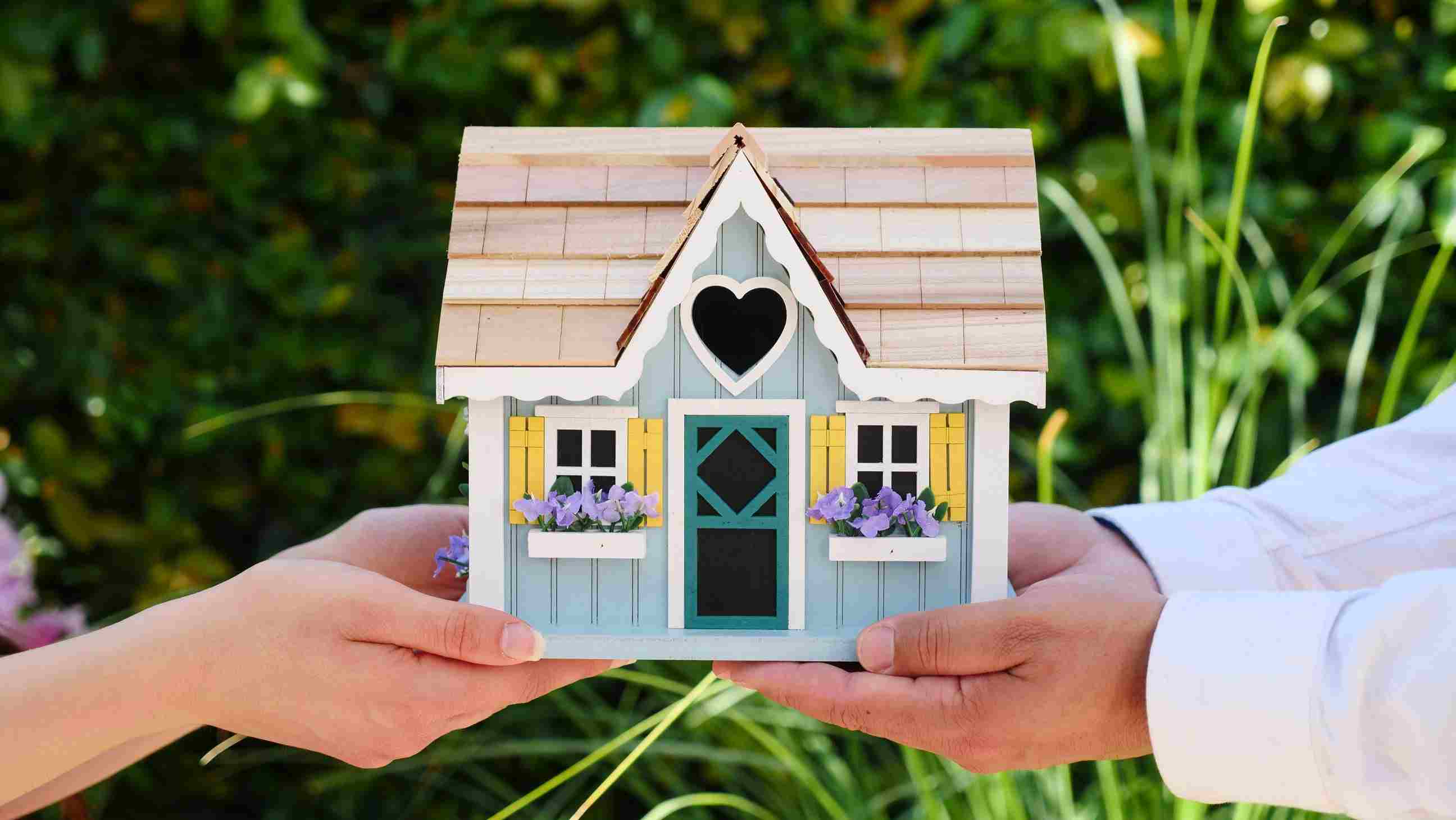 Two people hold a birdhouse between them. The birdhouse looks like a miniature house, with wooden roof tiles, a blue door, white windows, yellow window shutters, and purple flowers growing in windowsill planters. The entrance to the birdhouse is shaped like a heart and is centered above the blue door.