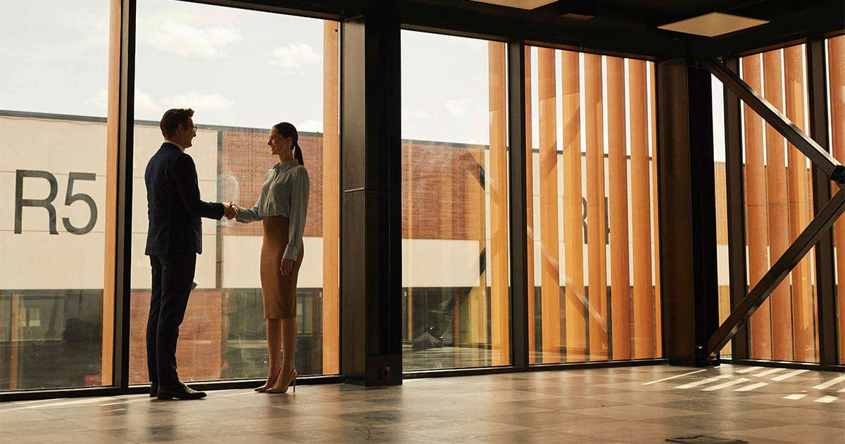 A man and woman stand by the window in an office building while they shake hands and smile at each other.