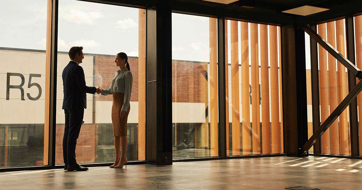 A man and woman stand by the window in an office building while they shake hands and smile at each other. 