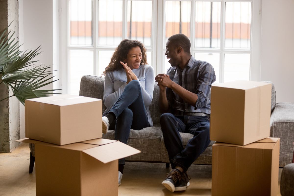 An excited couple sits on a couch surrounded by cardboard moving boxes. They clap their hands and smile enthusiastically.  