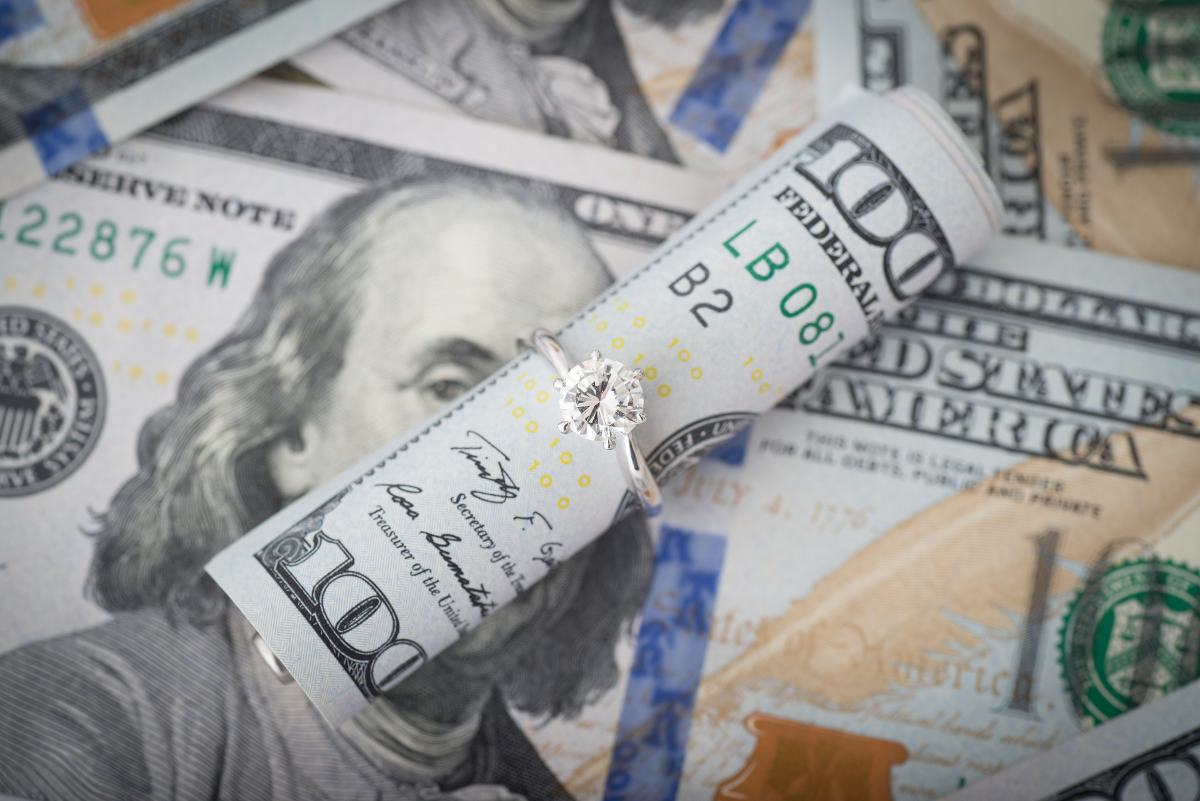 A diamond engagement ring rests on top of a pile of hundred dollar bills.