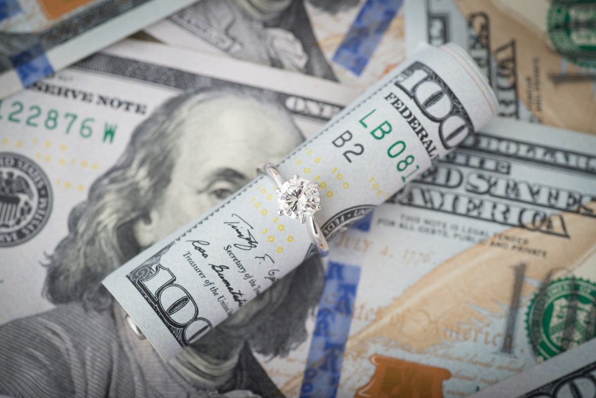 A diamond engagement ring rests on top of a pile of hundred dollar bills. 