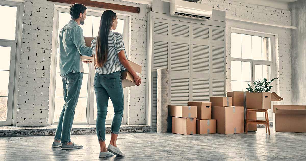 A couple carries moving boxes into a mostly empty room with white brick walls and several bright windows.