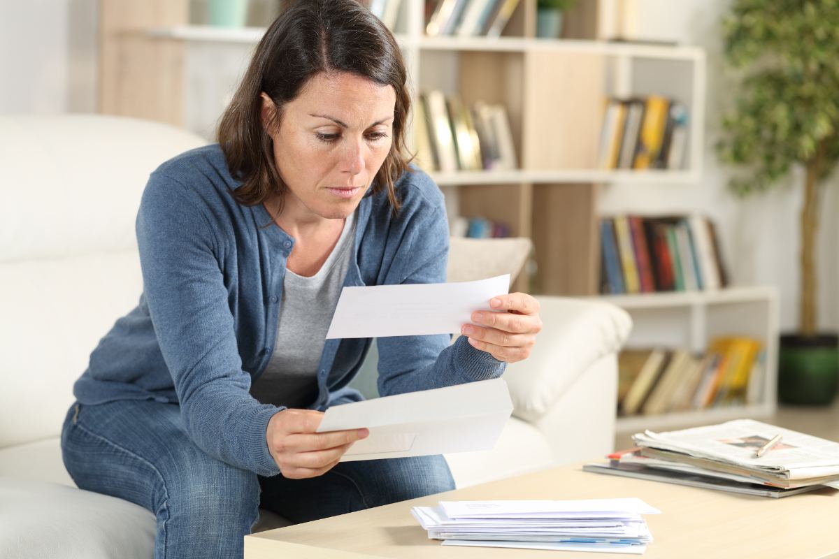 A woman with a somber expression sits on a couch and opens a letter.