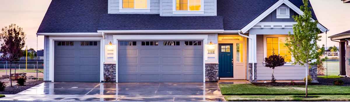 A large blue house with two front-facing garages. Yellow light shines through the living room and upstairs windows. 